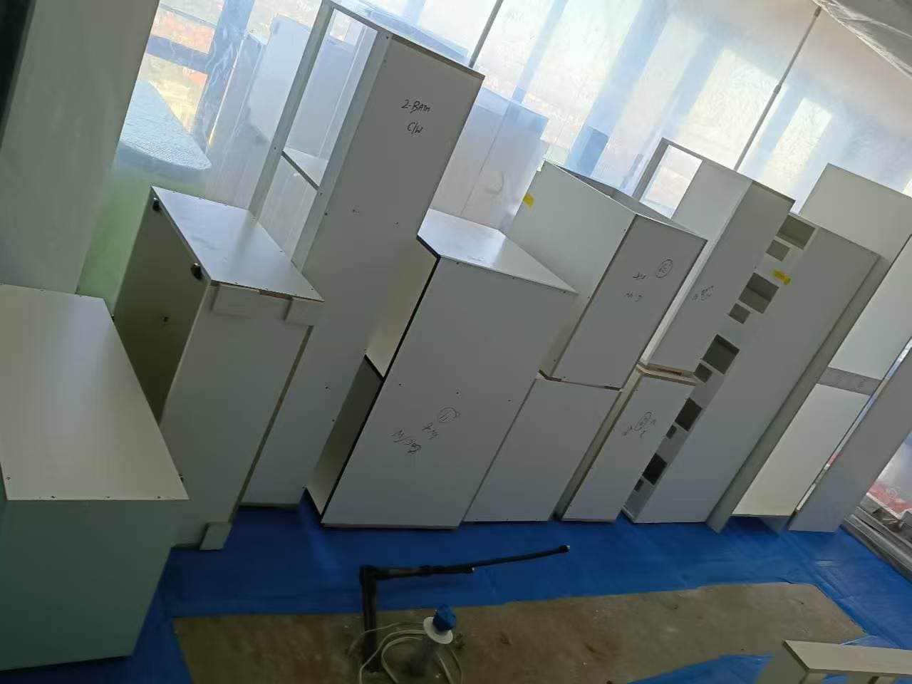 White modular cabinets standing on a blue tarp in a room with plastic-covered windows.