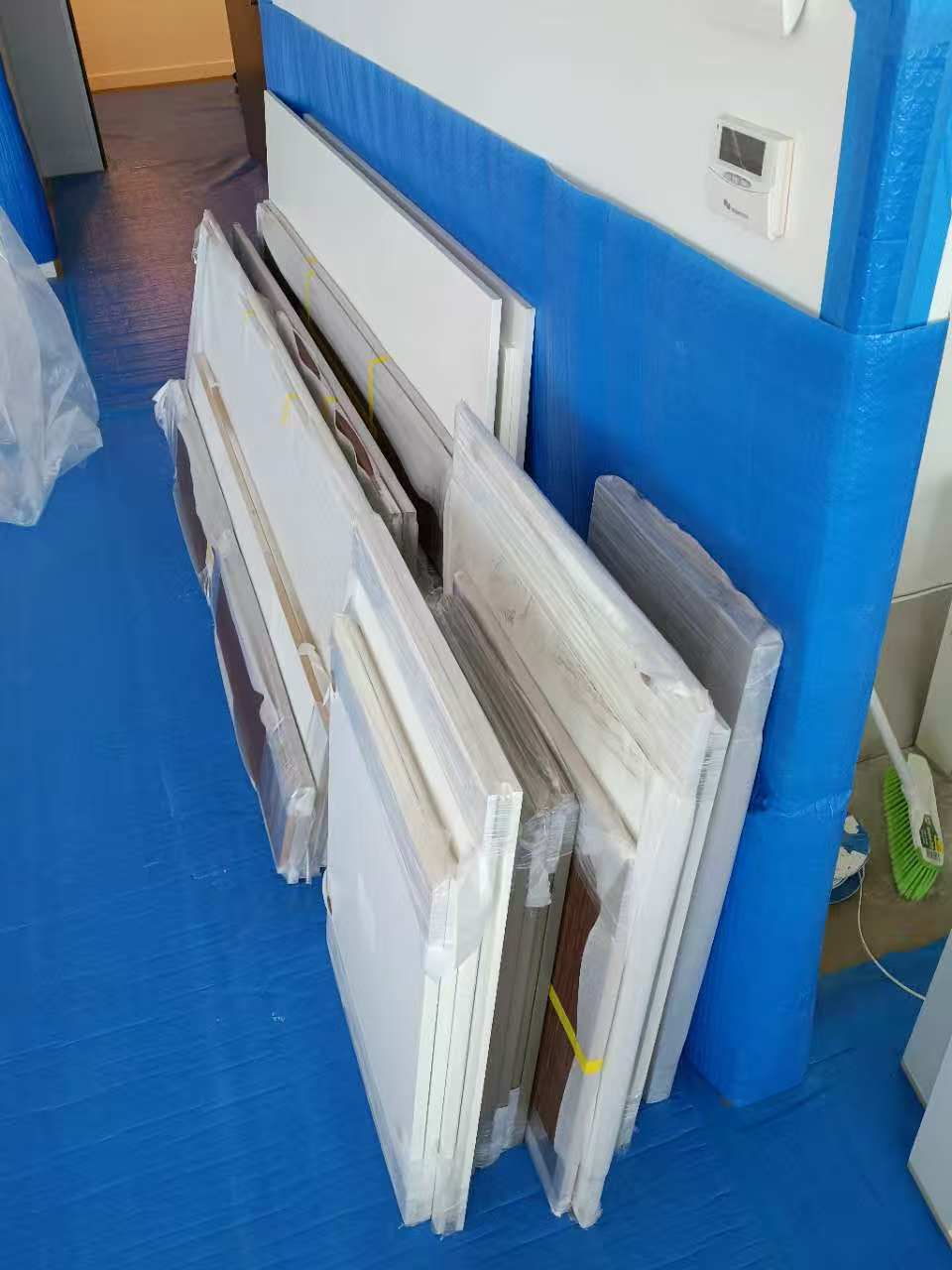 Plastic-wrapped panels leaning against a wall protected by blue sheeting in a construction zone.