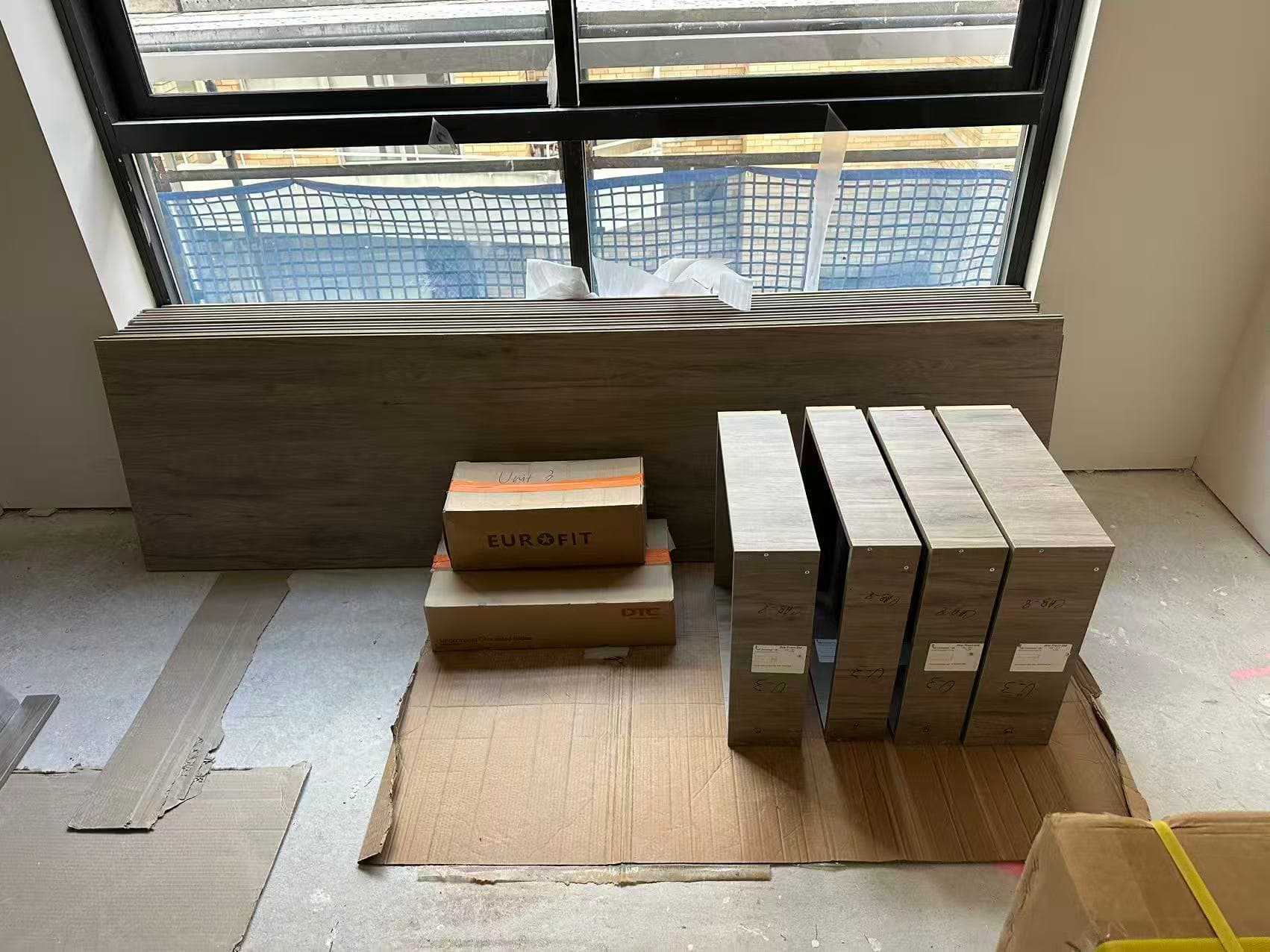 Stacked wood panels, cardboard boxes, and assembled cabinet frames on a construction site floor.