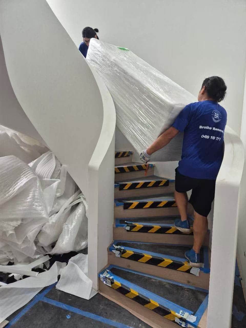 Two movers carry a large, plastic-wrapped piece of furniture up a curved indoor staircase.