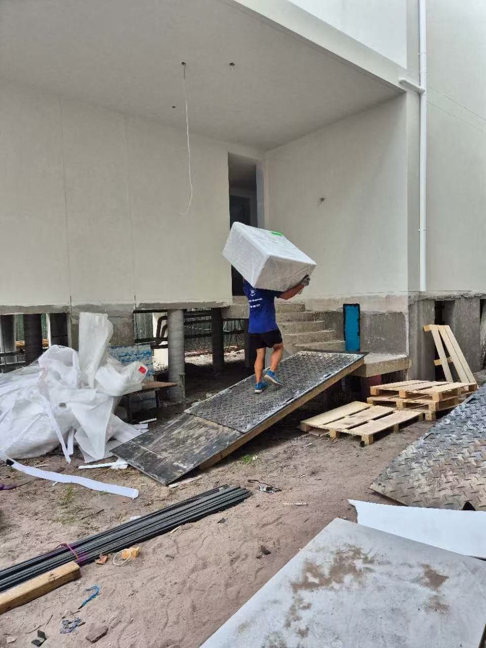 Worker carrying a large white box on their shoulder up a ramp into a building.