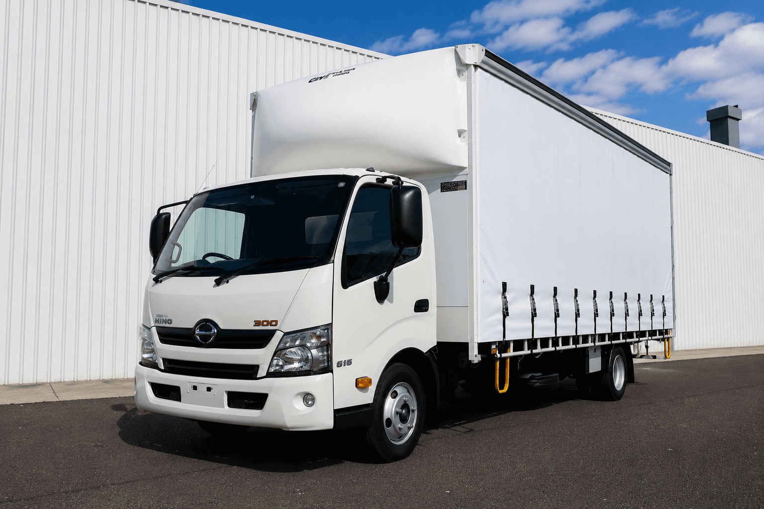 White Hino 300 series curtain-side truck parked outside a white corrugated metal warehouse.